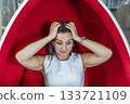 A stressed young Caucasian woman with short brown hair sits in a red egg chair, holding her head in her hands during a work break in an office environment. 133721109