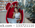 young Caucasian woman with brown hair sits in a red egg chair, using a laptop and talking on a phone in a modern coworking space. 133721110