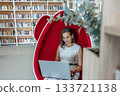 young woman with brown hair works on a laptop in a modern coworking space. She sits in a stylish red egg chair surrounded by bookshelves. 133721138