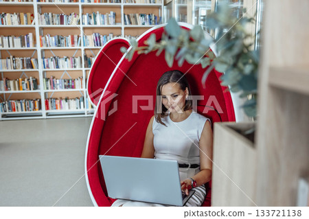 young woman with brown hair works on a laptop in a modern coworking space. She sits in a stylish red egg chair surrounded by bookshelves. young woman with brown hair works on a laptop in a modern coworking space. She sits in a stylish red egg chair surrounded by bookshelves. 133721138