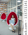 young Caucasian woman with brown hair sits in a red egg chair, working on a laptop in a modern coworking space with bookshelves in the background. 133721141