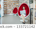 young Caucasian woman with brown hair works on a laptop in a modern coworking space. She sits in a stylish red egg chair surrounded by bookshelves. 133721142