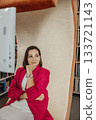 A young Caucasian woman with brown hair sits thoughtfully in a modern coworking space. She wears a red blazer and has a relaxed posture, surrounded by bookshelves. 133721143