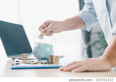 Concept of saving money and financial planning. A businesswoman puts coin into blue piggy bank on office desk with documents and laptop, symbolizing investment, budget management and future growth. 133721152