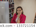 A young Caucasian woman with brown hair sits in a coworking space. She wears a red blazer and looks thoughtfully at the camera. Bookshelves are visible in the background. 133721154