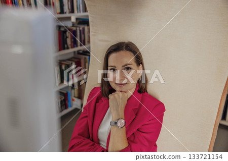 A young Caucasian woman with brown hair sits in a coworking space. She wears a red blazer and looks thoughtfully at the camera. Bookshelves are visible in the background. 133721154