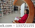 A young Hispanic woman with brown hair sits in a modern orange chair, working on a laptop in a coworking space filled with bookshelves. 133721155