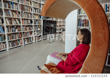 A young Hispanic woman with brown hair sits in a modern orange chair, working on a laptop in a coworking space filled with bookshelves. A young Hispanic woman with brown hair sits in a modern orange chair, working on a laptop in a coworking space filled with bookshelves. 133721155