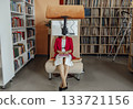 Unrecognizable woman sits in a modern coworking space. She wears a red blazer and white dress, typing on a laptop. Bookshelves are in the background. 133721156