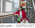 woman with short brown hair sits on an couch in a library, with a book. Bookshelves filled with books are visible in the background. 133721157