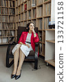 A young Caucasian woman with long dark hair sits in a modern chair, wearing a red blazer and white skirt, listening to music in a coworking space filled with vinyl records. 133721158