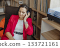 A young Caucasian woman with brown hair wears headphones while relaxing in a coworking space. A record player and vinyl records are visible in the background. 133721161