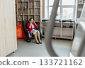 A young Asian woman in a red blazer sits in a modern coworking space. She is on the phone, surrounded by bookshelves and a relaxed atmosphere. 133721162