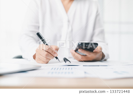 Concept of business planning and productivity. A businessman writes notes in a notebook while holding a smartphone, working with reports and laptop for financial analysis at modern office desk. 133721448