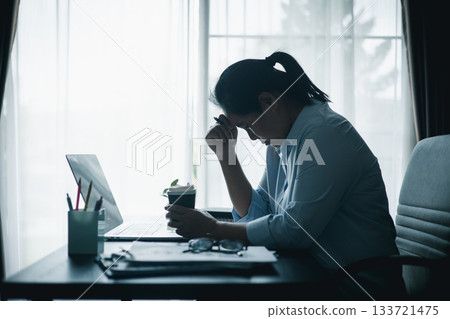 Stressed Asian woman sitting at desk with laptop, holding coffee cup and bowing head in prayer, symbolizing workplace stress, anxiety and finding hope or faith during difficult times. Stressed Asian woman sitting at desk with laptop, holding coffee cup and bowing head in prayer, symbolizing workplace stress, anxiety and finding hope or faith during difficult times. 133721475