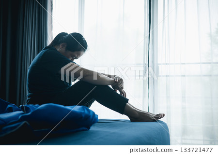 A stressed woman sitting alone on bed in dark room, holding her head down, symbolizing depression, anxiety, hopelessness, loneliness, mental health struggle, burnout and emotional pain. A stressed woman sitting alone on bed in dark room, holding her head down, symbolizing depression, anxiety, hopelessness, loneliness, mental health struggle, burnout and emotional pain. 133721477