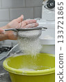 Hand using sieve to sift flour into yellow bowl. Household baking process symbolizing ingredient preparation, dough making. Closeup of flour passing through mesh sieve. vertical 133721865