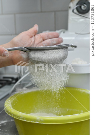 Hand using sieve to sift flour into yellow bowl. Household baking process symbolizing ingredient preparation, dough making. Closeup of flour passing through mesh sieve. vertical 133721865