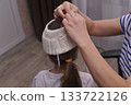 Little girl with ponytail is trying on a knitted hat made by her mother. Child stands still as mother adjusts a knitted hat. 133722126