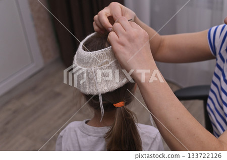 Little girl with ponytail is trying on a knitted hat made by her mother. Child stands still as mother adjusts a knitted hat. 133722126