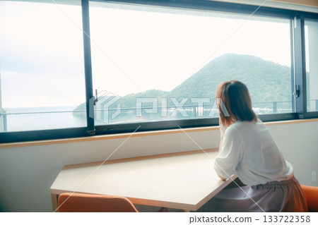 A scene of a woman in her twenties looking at the ocean and mountain scenery in a hotel room 133722358