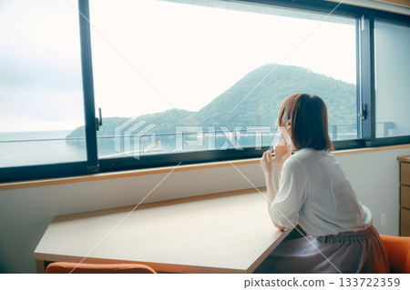 一位二十多歲的女子在飯店房間裡眺望大海和山脈的景色。 133722359