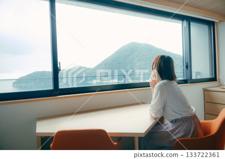 A scene of a woman in her twenties looking at the ocean and mountain scenery in a hotel room 133722361