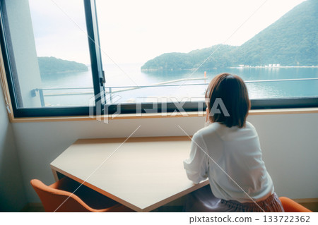 一位二十多歲的女子在飯店房間裡眺望大海和山脈的景色。 133722362