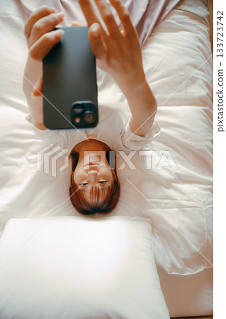 A scene of a woman in her 20s taking a selfie with her smartphone on a hotel bed 133723742