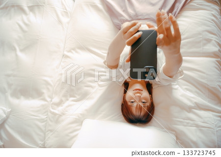 一位二十多歲的女子在飯店床上用智慧型手機自拍的場景 133723745