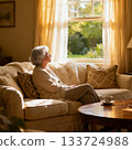 A gray-haired senior woman looking out the window from the sofa in her room 133724988
