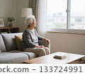 An elderly woman with gray hair sitting on a sofa in a bright room looking out the window 133724991