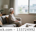 An elderly woman with gray hair sitting on a sofa in a bright room looking out the window 133724992