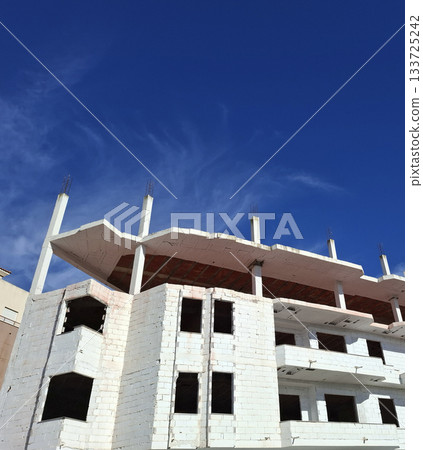A white brick house stands tall under construction, with its framework visible against a bright blue sky. The scene captures the progress of modern housing development. 133725242