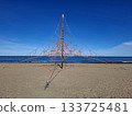 A bright children's climbing frame stands on the sandy beach, where kids can play and explore. The calm sea and clear sky create a perfect setting for a day of outdoor fun. 133725481