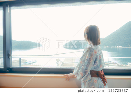 一位二十多歲的女子身穿浴衣,正從飯店窗戶向外眺望風景。 一位二十多歲的女子身穿浴衣,正從飯店窗戶向外眺望風景。 133725887