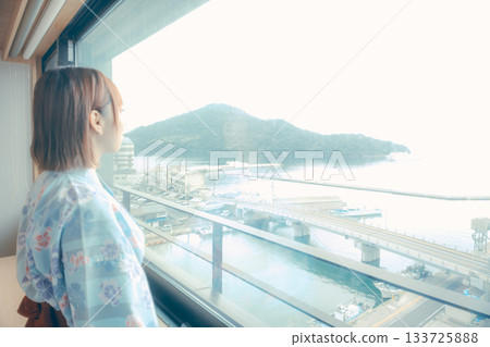 一位二十多歲的女子身穿浴衣，正從飯店窗戶向外眺望風景。 133725888