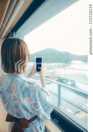一位二十多歲的女子身穿浴衣，正從飯店窗戶向外眺望風景。 133726219