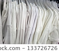 In a busy clothing store, numerous white cotton t shirts are displayed for sale. Shoppers browse while enjoying the bright afternoon light filtering through the windows. 133726726