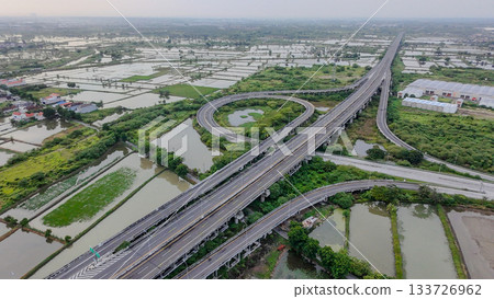 Aerial drone shot of a highway interchange overpass amidst lush green rice paddies and a river, showcasing infrastructure in nature. 133726962