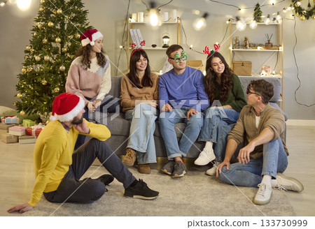 Young friends laughing and chatting by Christmas tree at home, cozy holiday vibes 133730999
