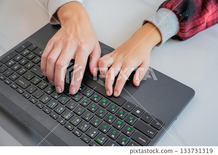 Close-up of adult and child hands typing together on a laptop. Only hands and keyboard visible. Concept of teaching, guidance, and digital learning. Close-up of adult and child hands typing together on a laptop. Only hands and keyboard visible. Concept of teaching, guidance, and digital learning. 133731319