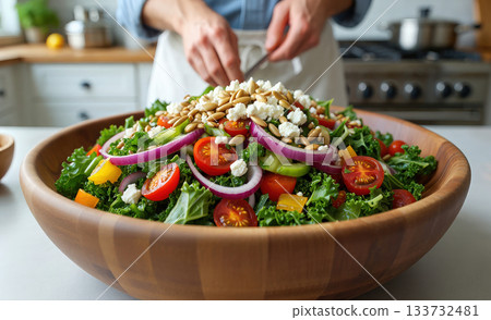 Close-up platter with a fresh salad on a wooden table and a person in background, chopping vegetables on a cutting board in a bright kitchen. Healthy eating and home cooking 133732481