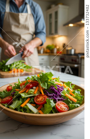 Close-up platter with a fresh salad on a wooden table and a person in background, chopping vegetables on a cutting board in a bright kitchen. Healthy eating and home cooking 133732482