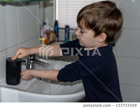 A preschooler in dark clothing stands at a bathroom sink, washing his hands using a black soap dispenser. The image emphasizes the importance of hygiene, teaching children cleanliness, and maintaining 133733509
