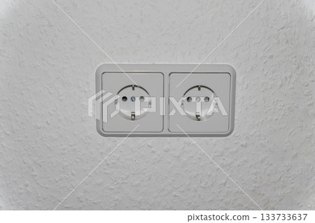 A double electrical outlet (Schuko type) in white or light gray is centered on a plain light-colored wall. The image focuses on themes of electricity, renovation, construction, home electrical systems 133733637