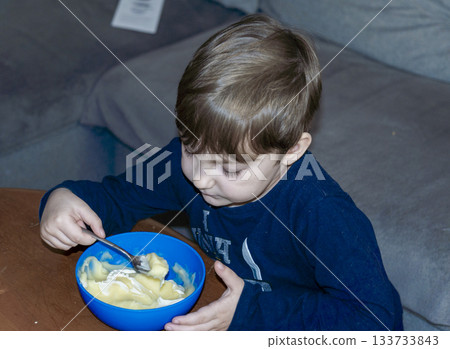 Photo from above. A boy in dark clothing sits at home, intently eating from a deep blue bowl with a spoon. The image reflects a daily routine, breakfast or snack time, and the child's focus on food. 133733843