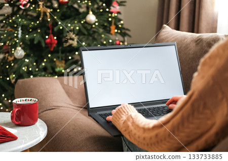 Woman in warm sweater is using laptop with blank white screen mock-up, sitting on sofa next decorated Christmas tree. Holiday online shopping, cozy winter digital lifestyle Woman in warm sweater is using laptop with blank white screen mock-up, sitting on sofa next decorated Christmas tree. Holiday online shopping, cozy winter digital lifestyle 133733885