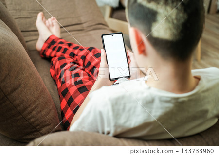 Boy wearing cozy pajama is lying comfortably on sofa holding smartphone with blank white screen, conveying relaxed holiday leisure and digital connection at home Boy wearing cozy pajama is lying comfortably on sofa holding smartphone with blank white screen, conveying relaxed holiday leisure and digital connection at home 133733960