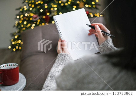 Over-the-shoulder view of person in sweater writing in blank spiral notebook with pen with blurry Christmas tree and lights in background. Goals, wish list, resolutions, planning, to-do list, mockup 133733962